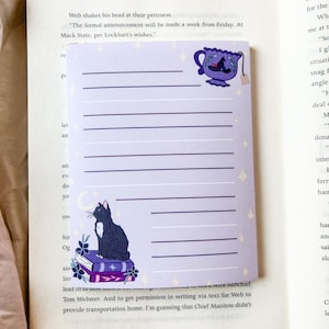 May include: A lined notepad with a purple and white design. The notepad features a black cat sitting on books, a teacup with a witch hat, and star and moon accents. The notepad is open on a book.