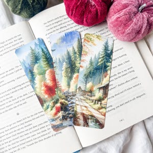 May include: Three landscape-themed bookmarks with watercolour illustrations of forests, streams, and cabins. The bookmarks are on top of an open book with text. The colour palette includes greens, reds, and browns.