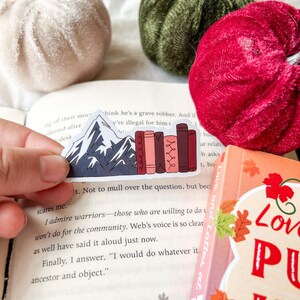 May include: A sticker featuring a mountain range and a row of books in shades of red and pink. The sticker is placed on an open book. Decorative velvet pumpkins in cream, green, and red are in the background.