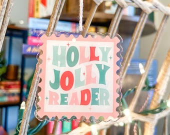 Könnte beinhalten: Ein klarer Acryl-Weihnachtsschmuck mit einem rosa, gezackten Rand. Der Schmuck zeigt die Worte "HOLLY JOLLY READER" in einer Retro-Schriftart mit roten, türkisfarbenen und rosa Farben. Ein rosa Faden ist befestigt.