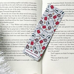 May include: A gray and white bookmark with a Christmas theme. The bookmark features a variety of festive designs, including candy canes, gingerbread men, ornaments, and books. The text "The JustinspiredShop" is printed on the bookmark.