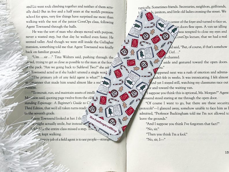 May include: A gray and white bookmark with a Christmas theme. The bookmark features a variety of festive designs, including candy canes, gingerbread men, ornaments, and books. The text "The JustinspiredShop" is printed on the bookmark.