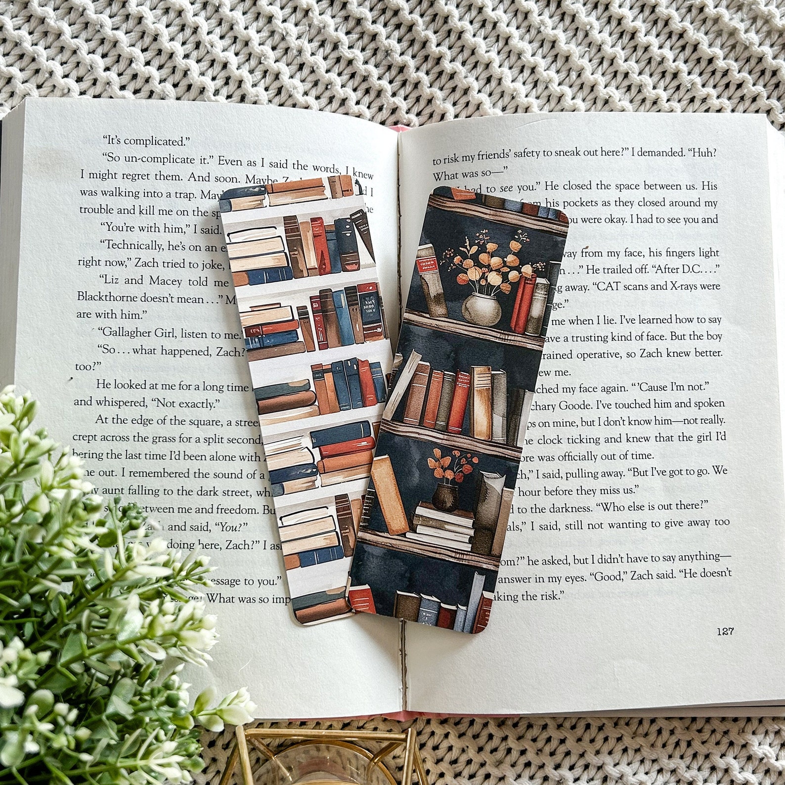 Two bookmarks lay on the pages of an opened book. The two bookmarks depict illustrations of bookshelves filled with books and plants. They are illustrated in warm hues and have navy undertones.