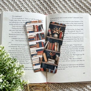 May include: Two bookmarks featuring a bookshelf design. The bookmarks are made of paper and have a watercolour look. One bookmark has a dark blue background with a bookshelf filled with books in shades of red, orange, and brown. The other bookmark has a light blue background with a bookshelf filled with books in shades of red, orange, and brown. Both bookmarks have a floral design in the bottom right corner.