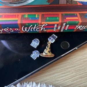 May include: A gold-coloured witch hat charm with a crescent moon and star design, dangling from a clear earring back. Two additional clear earring backs are also present. The items are on a black surface.