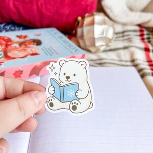 May include: A cartoon sticker of a white polar bear reading a blue book with a snowflake. The sticker is held in front of a blank notebook. Other items in the background include a book and Christmas ornaments.