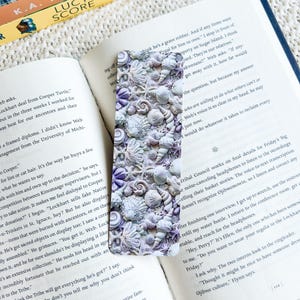 May include: A rectangular bookmark with a detailed relief of seashells in white, grey, and purple. The bookmark is on an open book.