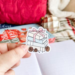 May include: A sticker showing a stack of books tied with a pink bow. The books are white and light blue, with small pinecones and stars at the base. The sticker is held over an open notebook.