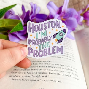 May include: A sticker with the text "Houston I'm Probably The Problem" in purple and teal, with a rocket ship graphic. The sticker is held in front of a book and purple flowers.