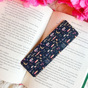 May include: A dark blue bookmark featuring a pattern of potion bottles, candles, moons, and leaves. The bookmark rests on an open book. The bookmark is rectangular in shape.