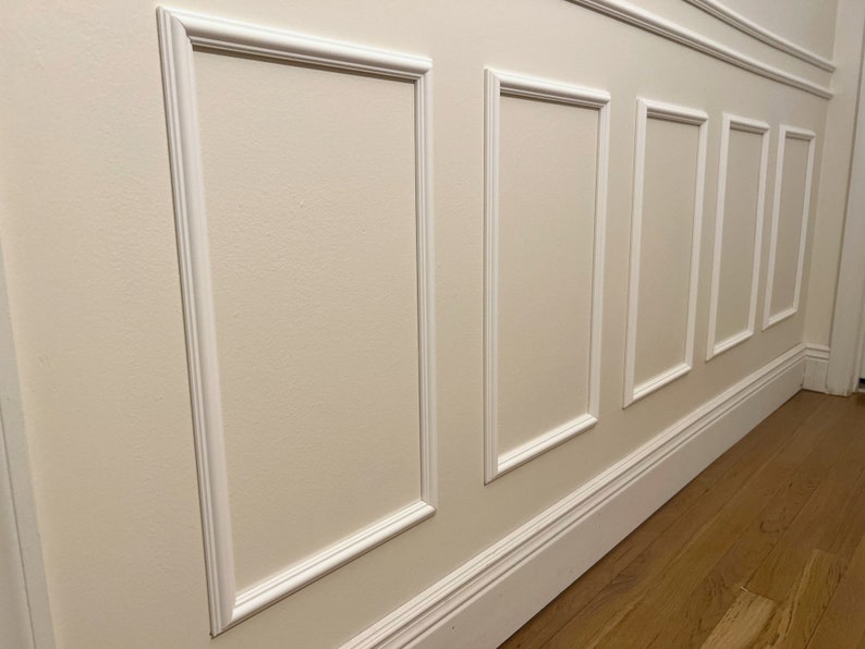 May include: A section of wall with decorative white molding. The wall is painted a light beige color, and the molding creates rectangular panels. The floor is made of wooden planks.