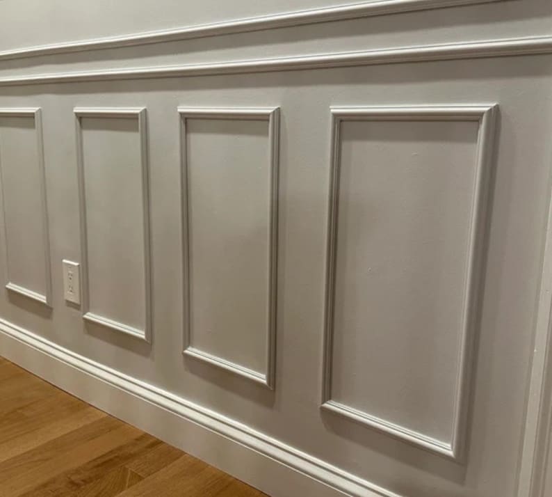 May include: A section of a wall with decorative molding. The wall is painted a light gray color and features rectangular panels framed by white molding. A power outlet is visible on the wall. The floor is made of wood.