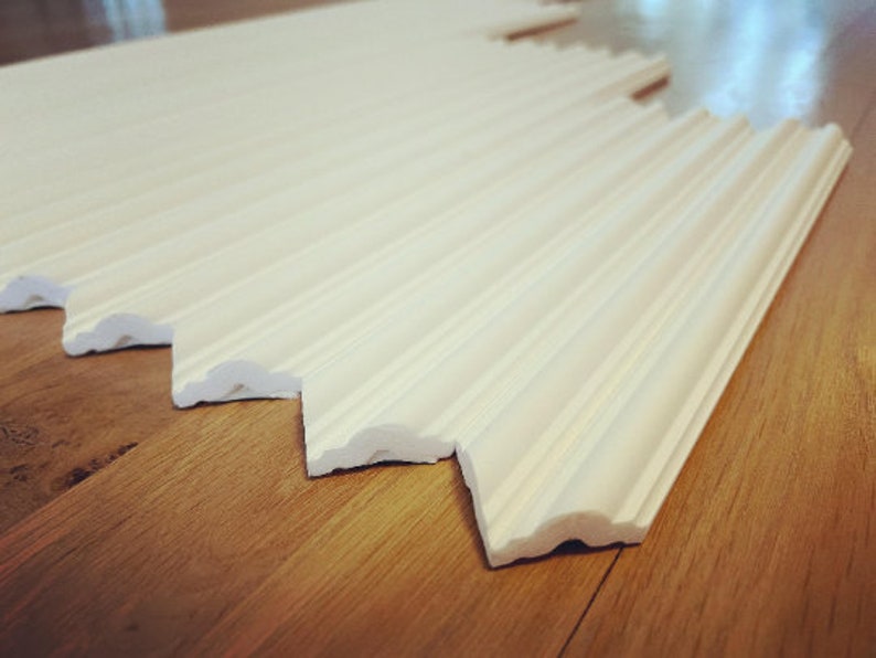 May include: Several sections of white crown molding are arranged diagonally on a wooden floor. The molding has a decorative, ridged design and a scalloped edge. The pieces are cut at an angle.