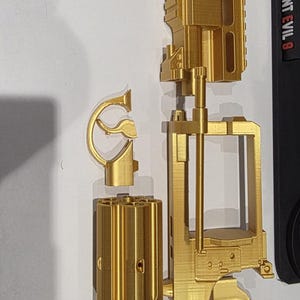 May include: A disassembled gold-coloured replica gun, possibly 3D-printed, with components like the barrel, grip, and cylinder. The grip has a decorative design. The gun is shown against a white background.