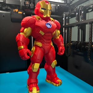 Op de afbeelding: Een 3D-geprint figuur van een personage in een rood en goudkleurig harnas met een helm, borstplaat en beweegbare ledematen. Het figuur heeft een blauw zeshoekig detail op de borst en staat op een vlak, felblauw oppervlak.