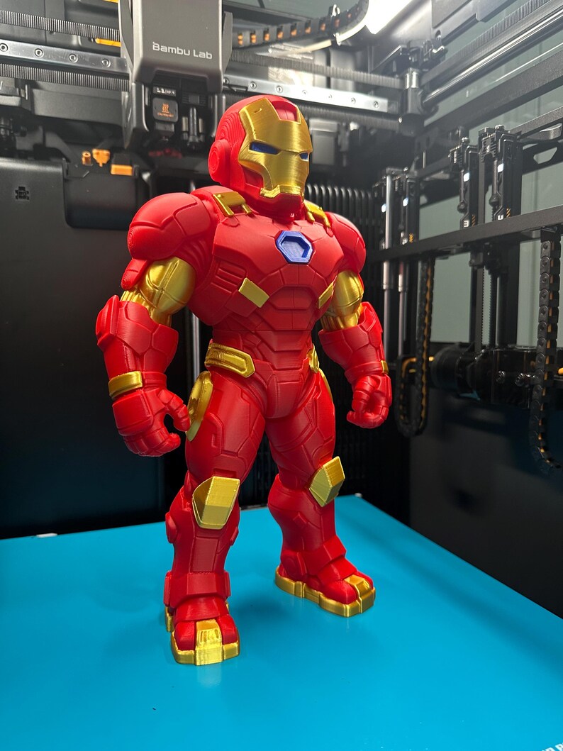 Op de afbeelding: Een 3D-geprint figuur van een personage in een rood en goudkleurig harnas met een helm, borstplaat en beweegbare ledematen. Het figuur heeft een blauw zeshoekig detail op de borst en staat op een vlak, felblauw oppervlak.