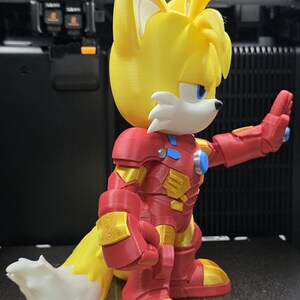Op de afbeelding: Een 3D-geprint figuur van een gele vos met witte accenten, gekleed in een rood en goudkleurig Iron Man-pak. Het figuur heeft blauwe ogen en een witgepunte staart. Het figuur staat voor een 3D-printer.