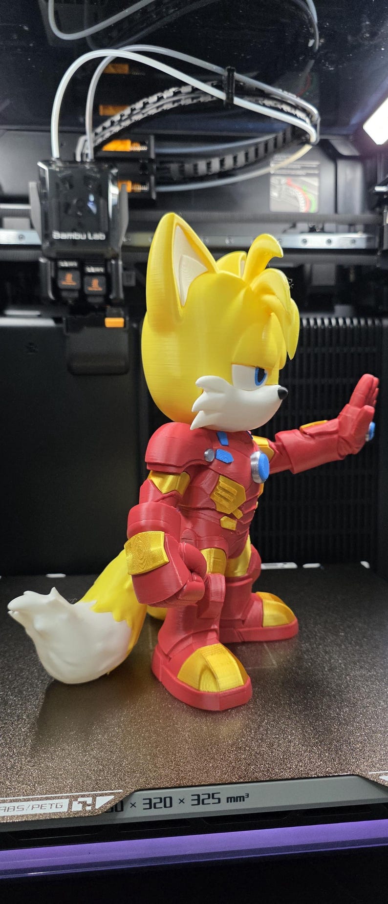 Op de afbeelding: Een 3D-geprint figuur van een gele vos met witte accenten, gekleed in een rood en goudkleurig Iron Man-pak. Het figuur heeft blauwe ogen en een witgepunte staart. Het figuur staat voor een 3D-printer.