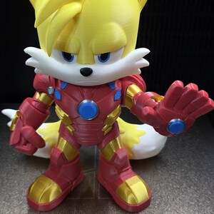 Op de afbeelding: Een 3D-geprint figuur van Tails uit Sonic the Hedgehog, gekleed in een Iron Man-pak. Het figuur is geel, rood en goud, met blauwe accenten. Het figuur staat op een 3D-printerbed.