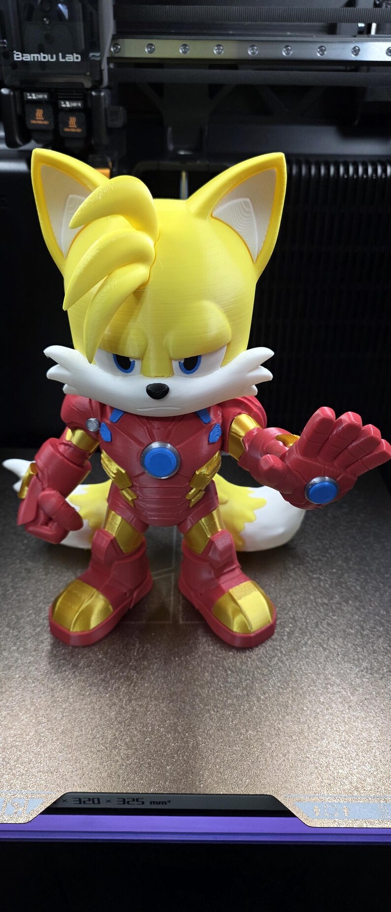 Op de afbeelding: Een 3D-geprint figuur van Tails uit Sonic the Hedgehog, gekleed in een Iron Man-pak. Het figuur is geel, rood en goud, met blauwe accenten. Het figuur staat op een 3D-printerbed.