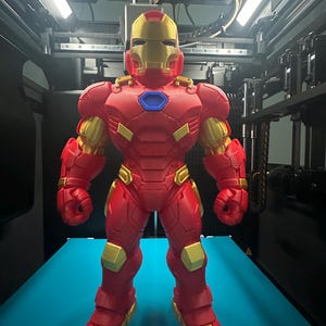 Op de afbeelding: Een 3D-geprint Iron Man-figuur met een rood en goudkleurig harnas met een metallic afwerking. Het personage heeft een opvallende gouden helm, een blauw zeshoekig borststuk en gearticuleerde beplating over de romp, armen en benen.