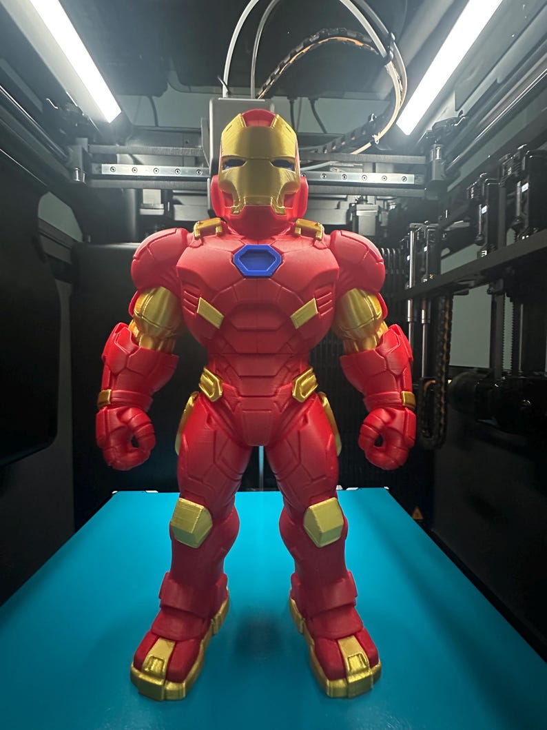 Op de afbeelding: Een 3D-geprint Iron Man-figuur met een rood en goudkleurig harnas met een metallic afwerking. Het personage heeft een opvallende gouden helm, een blauw zeshoekig borststuk en gearticuleerde beplating over de romp, armen en benen.