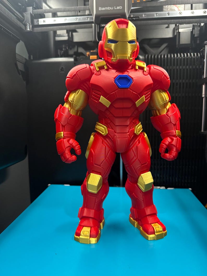 Op de afbeelding: Een 3D-geprinte Iron Man-actiefiguur met een rood en goudkleurig pak en een blauwe arc-reactor op de borst. Het figuur heeft een goudkleurig masker, gedetailleerde bepantsering en staat in een neutrale houding met een robuust ontwerp.