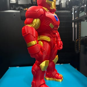 Op de afbeelding: Een 3D-geprint figuur in een rood en goudkleurig harnas. Het figuur heeft een metallic gouden gezichtsplaat, borstaccenten en gewrichtsdetails, met een felrood lichaam en een klein blauw cirkelvormig embleem in het midden van de borst.