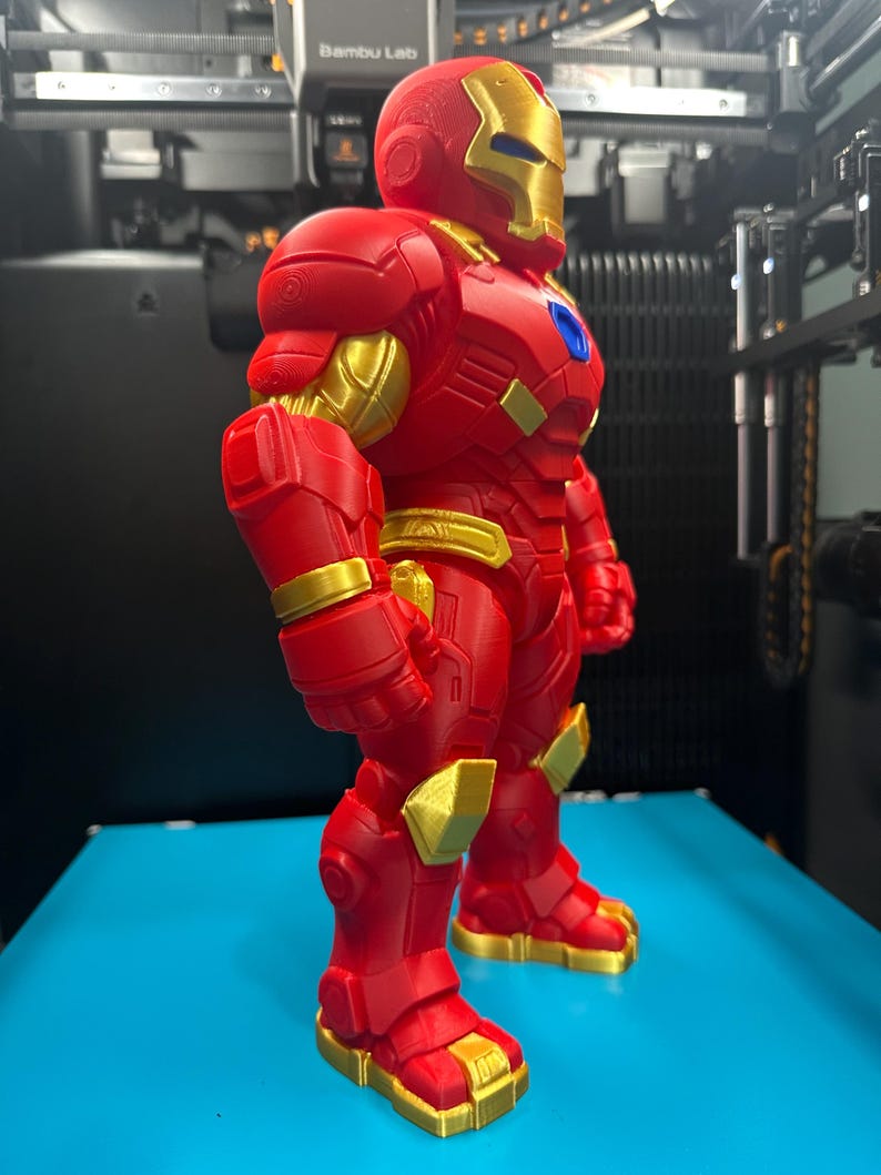 Op de afbeelding: Een 3D-geprint figuur in een rood en goudkleurig harnas. Het figuur heeft een metallic gouden gezichtsplaat, borstaccenten en gewrichtsdetails, met een felrood lichaam en een klein blauw cirkelvormig embleem in het midden van de borst.