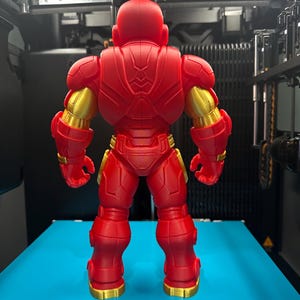 Op de afbeelding: Een 3D-geprinte actiefiguur van een robotachtig personage dat met de rug naar de kijker toe staat. Het figuur is voornamelijk matrood met metallic gouden accenten op de schouders, armen en voeten, met ingewikkelde mechanische details.