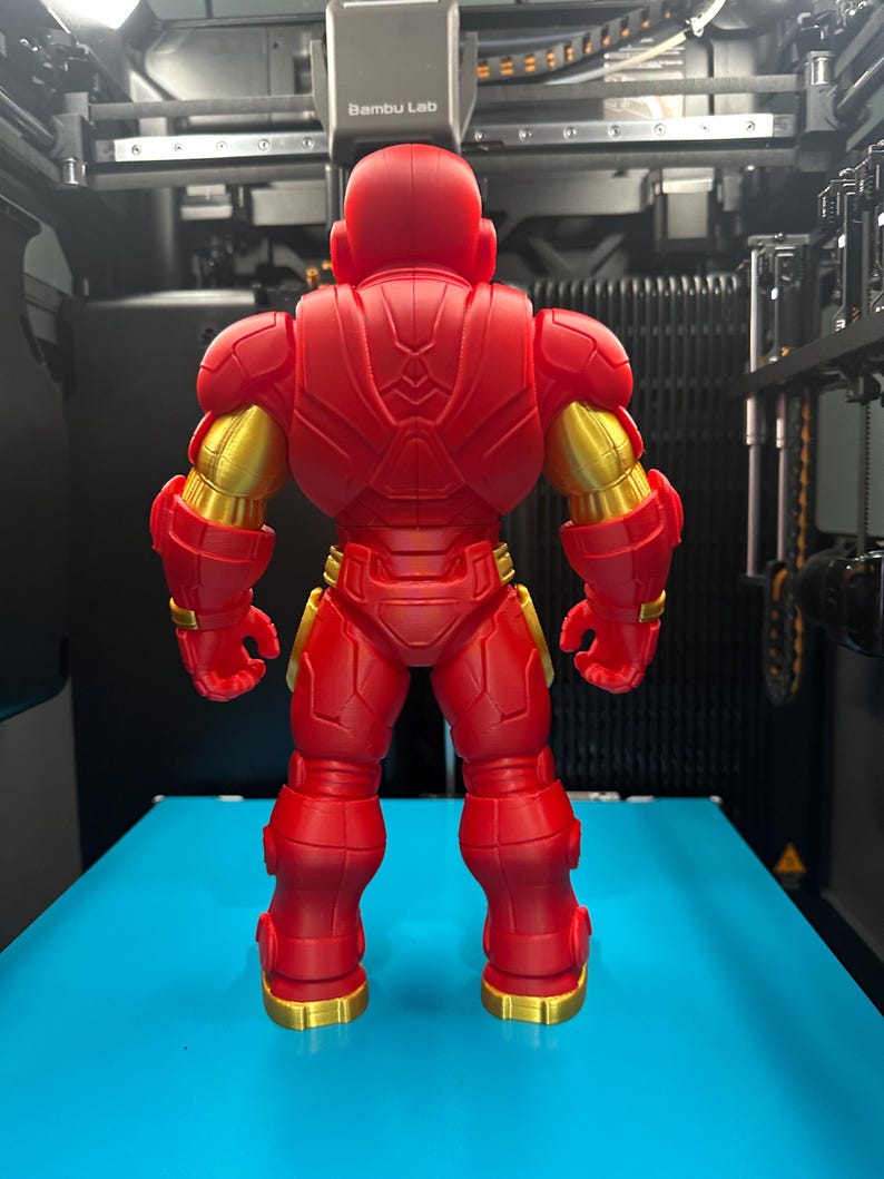 Op de afbeelding: Een 3D-geprinte actiefiguur van een robotachtig personage dat met de rug naar de kijker toe staat. Het figuur is voornamelijk matrood met metallic gouden accenten op de schouders, armen en voeten, met ingewikkelde mechanische details.