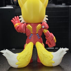 Op de afbeelding: Een 3D-geprint beeldje van een vos-personage in een rood en goudkleurig Iron Man-achtig pak. De vos heeft gele oren en een gele kop, met twee grote, pluizige, geel en witte staarten. De figuur poseert met &eacute;&eacute;n hand omhoog.