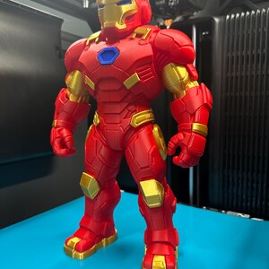 Op de afbeelding: Een staand Iron Man-actiefiguur met een matrode en metallic gouden kleurenschema. Het personage heeft een blauwe cirkelvormige arc-reactor op de borst, een gedetailleerd pantserpak met gesegmenteerde platen en een helm met een gouden vizier.