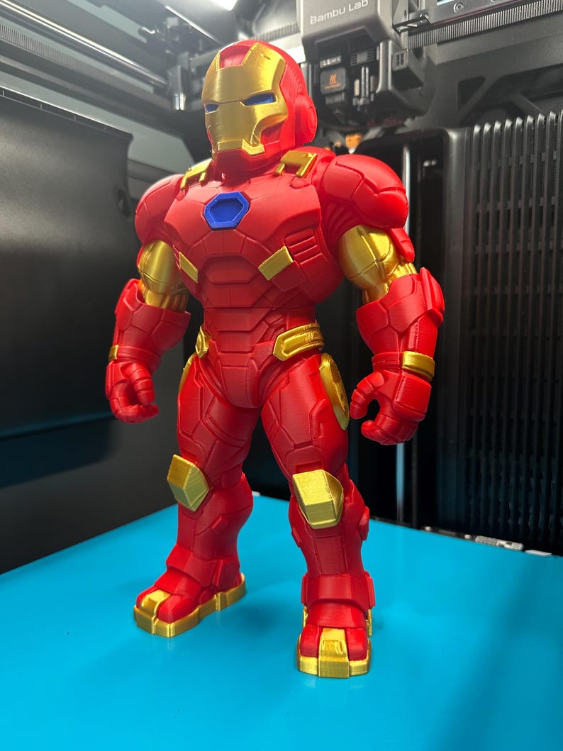 Op de afbeelding: Een staand Iron Man-actiefiguur met een matrode en metallic gouden kleurenschema. Het personage heeft een blauwe cirkelvormige arc-reactor op de borst, een gedetailleerd pantserpak met gesegmenteerde platen en een helm met een gouden vizier.