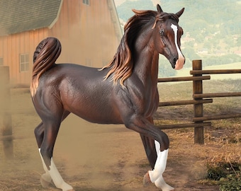 May include: A brown and white Arabian horse figurine with a long flowing mane and tail. The horse is standing on a dirt path with a wooden fence and a barn in the background.