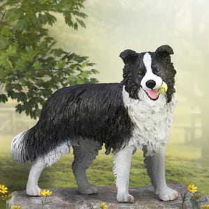 May include: A black and white border collie figurine with a playful expression, holding a green ball in its mouth. The figurine is standing on a gray rock with green grass and yellow flowers in the background.