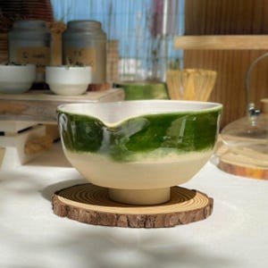 Può includere: Una ciotola per matcha in ceramica con smalto verde e crema, appoggiata su un sottobicchiere di legno. La ciotola ha un beccuccio per versare facilmente. Altri oggetti legati al tè sono visibili sullo sfondo, suggerendo una cerimonia del tè.