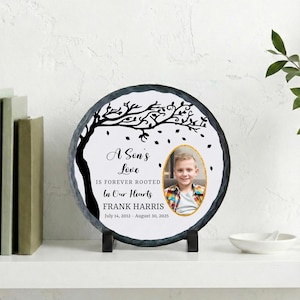 May include: A round, black slate memorial plaque with a white center featuring a tree silhouette and the text "A Son's Love Is Forever Rooted In Our Hearts." Includes a photo and the name Frank Harris with dates.