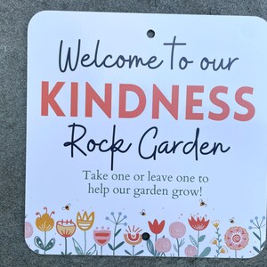 May include: A white square sign with rounded corners. The sign reads "Welcome to our KINDNESS Rock Garden" in black and coral lettering. The bottom of the sign features a floral design with bees.
