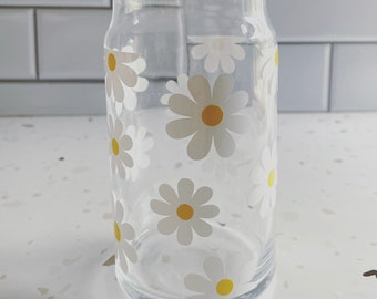 May include: A clear glass can with a daisy pattern. The daisies are white with yellow centers.