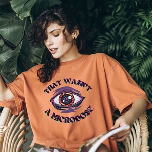 May include: A woman wearing an orange t-shirt with a graphic of an eye and the text "THAT WASN'T A MICRODOSE".