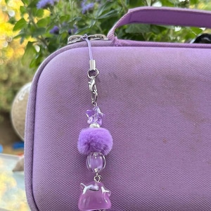 May include: A purple beaded phone charm featuring a small star-shaped bead, a fuzzy purple pom-pom, a clear round bead, a translucent purple ghost-shaped charm, and a miniature purple handheld game console charm with the word Win printed on it.