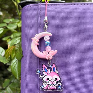 Puede incluir: Un charm rosa y blanco con un personaje de dibujos animados con detalles azules, una luna creciente rosa y cuentas. El charm está sujeto a un cordón rosa y cuelga sobre un fondo morado.