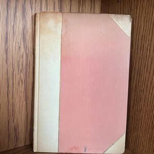 May include: A vintage book with a pink cover and a cream-colored spine. The book's cover has a slightly faded appearance, with a gold accent on the corner. The book is displayed on a wooden shelf.