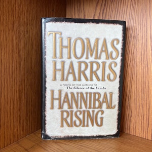 Puede incluir: Un libro de tapa dura titulado "Hannibal Rising" de Thomas Harris. El libro tiene una cubierta blanca y beige con letras doradas. También se puede ver el texto "A Novel by the Author of The Silence of the Lambs".