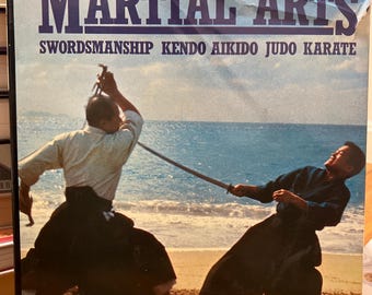 Op de afbeelding: Een boekomslag met de titel "The Martial Arts: Swordsmanship, Kendo, Aikido, Judo, Karate" met een foto van twee mensen die vechtsporten beoefenen op een strand. Het boek is van Michel Random.