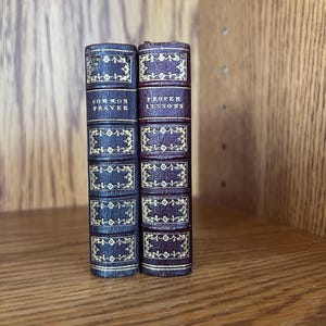 May include: Two small, antique leather-bound books standing upright. The left book is dark blue with gold-embossed floral patterns and the text "COMMON PRAYER". The right book is deep red with similar gold detailing and the text "PROPER LESSONS".