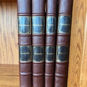 Könnte beinhalten: Vier antike Bücher mit braunen Lederrücken und goldener Schrift. Die Titel lauten "Shakespeare", "Komödien", "Historien", "Zweifelhaft" und "Tragödien". Die Bücher werden auf einem Holzregal ausgestellt.
