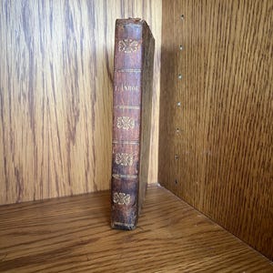 May include: A vintage book with a brown leather cover and gold lettering. The spine features decorative gold floral patterns and the title "Г. АНДРЕЕВ". The book is displayed on a wooden shelf.