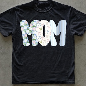 Könnte beinhalten: Ein schwarzes Kurzarm-T-Shirt mit dem Wort MOM in großen Buchstaben auf der Brust. Der Buchstabe M hat ein blaues Blumenmuster, das O ist mit kleinen Schleifen verziert und das letzte M zeigt ein vertikales blau-weißes Streifenmuster.